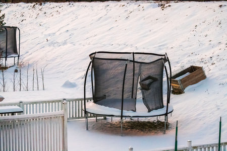 Trampoline with open entrance in safety net standing near white fence in snow-covered winter backyard. Sweden.の写真素材