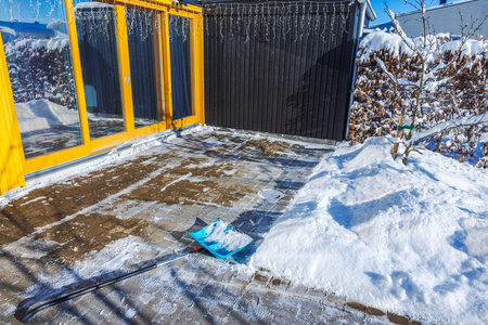 Blue snow shovel lying on cleared brick patio near panoramic windows of villa on sunny winter day. Sweden.の写真素材