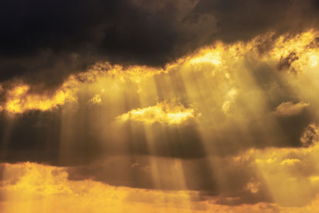 Dramatic golden sunlight breaking through dark storm clouds, creating radiant beams illuminating sky at sunset.の写真素材