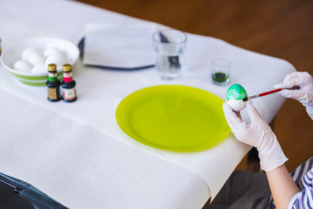 Child carefully painting Easter egg with brush while wearing gloves at decorated table with colors and raw eggs.の写真素材