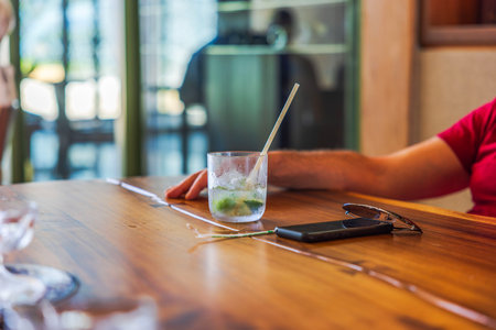Cold drink with ice, lime, and straw on wooden table near smartphone and sunglasses, with man resting arm nearby. Mexico.の写真素材