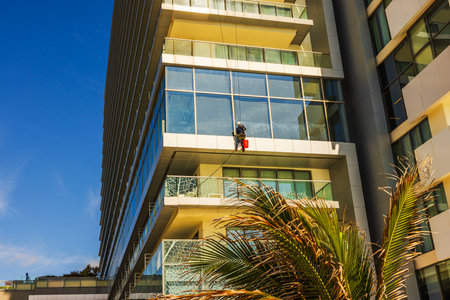 Worker suspended on ropes cleaning glass windows of modern hotel under blue sky with palm tree in foreground. Mexico. Cancun.の写真素材