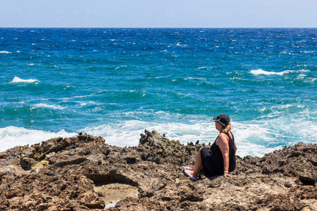Woman sitting on rocky shore looking at turquoise waves of Caribbean Sea under bright sunlight and clear blue sky. Aruba.の写真素材