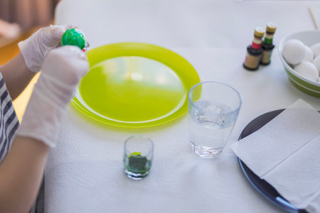 Person wearing gloves painting Easter egg green with brush on table set with dye, water glasses and white eggs. Sweden.の写真素材