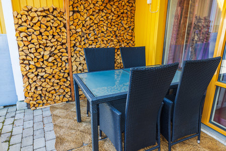Cozy patio of villa with wicker chairs, glass table, and stacked firewood by yellow wooden wall. Sweden.の写真素材
