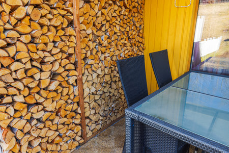 Stacked firewood beside glass table and black chairs on cozy outdoor patio with yellow wooden wall villa. Sweden.の写真素材