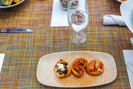 Three assorted bruschettas with toppings served on textured plate with glass of water on table.の写真素材