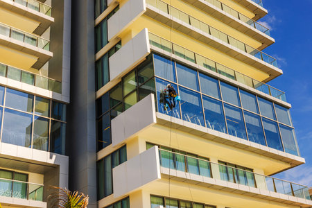 Worker suspended on ropes cleaning windows of modern hotel building with blue sky in Caribbean resort. Mexico. Cancun.の写真素材