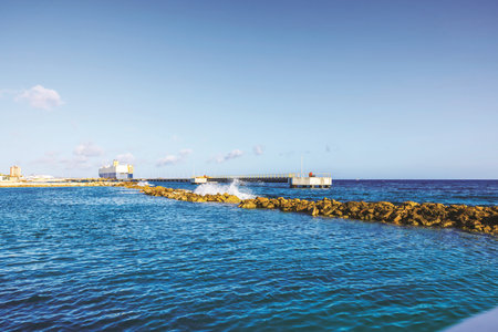 Waves crashing on stone breakwater near industrial pier and blue water of Caribbean Sea under clear sky. Curacao.の写真素材