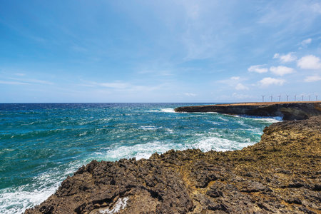 Rocky coastline with waves of Caribbean Sea hitting shore under sunny sky and wind turbines in background. Aruba.の写真素材
