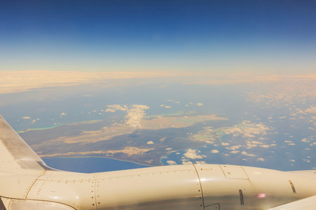 Airplane wing flying above clouds and coastline with bright blue sky on background during daytime...
