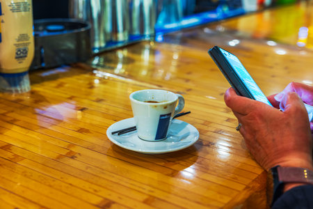 Close up view of coffee cup on wooden counter as woman uses smartphone inside airport cafe. Miami. USA. 11.24.2025.のeditorial素材