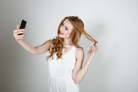 Beautiful girl with red hair and bright red lipstick smiling and looking at the camera on a light background makes the SELFの写真素材