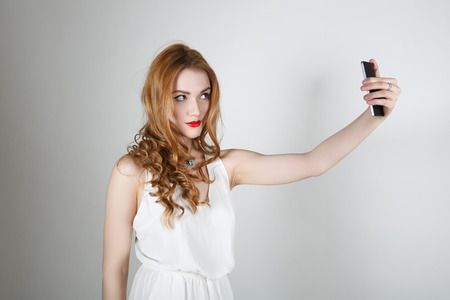 Beautiful girl with red hair and bright red lipstick smiling and looking at the camera on a light background makes the SELFの写真素材
