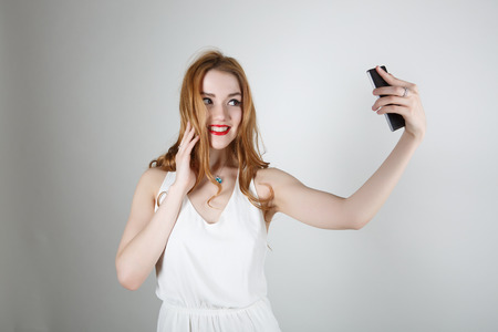 Beautiful girl with red hair and bright red lipstick smiling and looking at the camera on a light background makes the SELFの写真素材