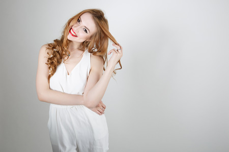 Cute girl with red hair and bright red lipstick smiling and looking at the camera on a light backgroundの写真素材