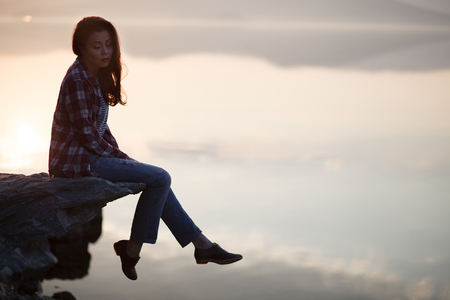 Woman sitting on a sand beach in front of sunset and ocean or lake in autumnの写真素材