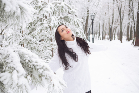 Outdoor portrait of cute young pretty attractive girl with long dark hair white warm sweater on natural snowy background in park. Outdoor winter photo in soft light. Lifestyleの写真素材