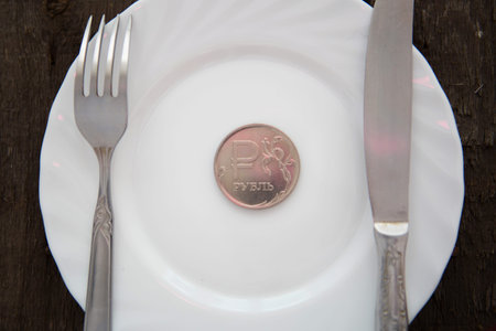 One ruble coin on a white plate next to fork and knife