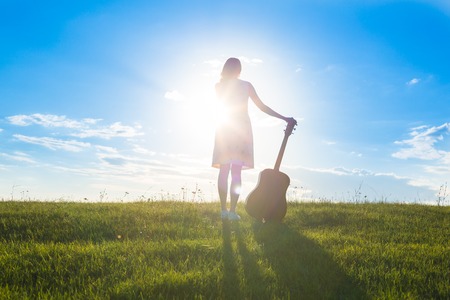 beautiful young blonde woman in dress holding the guitar on cloudy sunset or sunrise sky and green field background Copy Space for inscriptionの写真素材