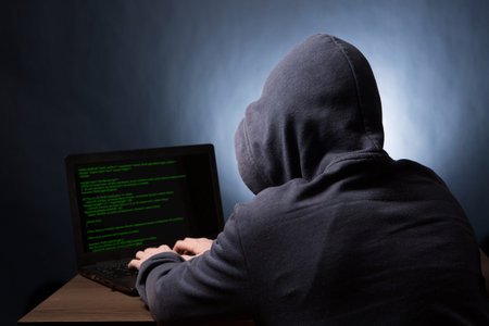 Hacker in hood with laptop initiating cyber attack. View from the back.