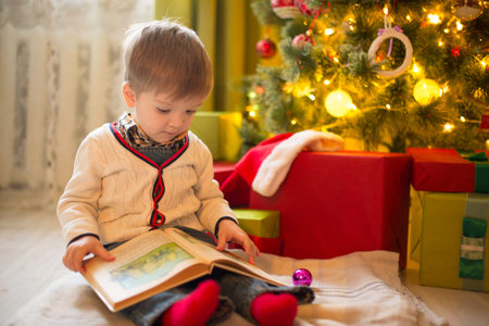 Happy little boy with book on Christmas Eve in decorated room. Sunny morning or day. Christmas or New Year holiday.の写真素材