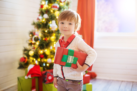 Little boy hold a gift box near a decorated Christmas tree at home.の写真素材