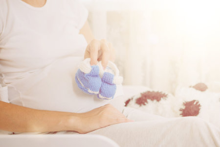 Pregnant girl in peach-colored negligee. Girl holding baby booties blue near pregnant belly.
