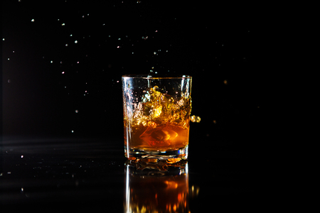 Ice cube falls into a glass with whiskey on black background. Fluid in motion. Droplets of spray.の写真素材