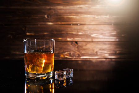 Glass of whiskey with ice cubes on a black table against wooden backgroundの写真素材