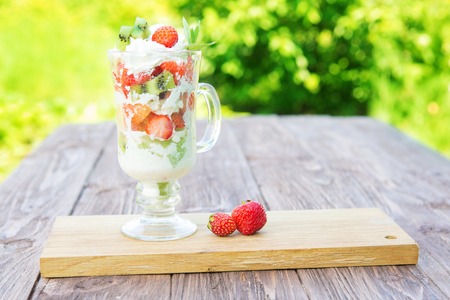 Strawberry layered dessert with fresh strawberries and cream cheese on wooden table over a green garden background.の写真素材