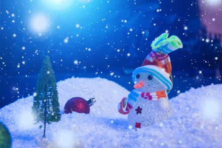 Snow Man with Christmas balls on snow over fir-tree, night sky and moon. Shallow depth of field. Christmas background. Fairy tale. Macro. Artificial magic dreamy world.の写真素材