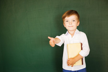 School child thumbs up and hold book over blackboard. Free text, copy space