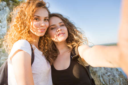 A world without borders. Stunning journey of two girls in the mountains. two attractive young girls travel together, do selfie on the phone against the sky and landscape in the mountains in the sunの写真素材