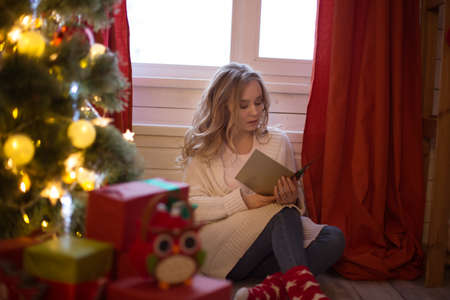 Young beautiful woman sitting on floor near christmas tree and reading christmas postcardの写真素材