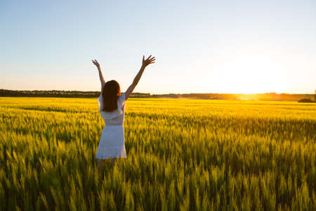 Beautiful caucasian woman in white dress jumping up with raised hands outdoor. Field with yellow wheat aroundの写真素材