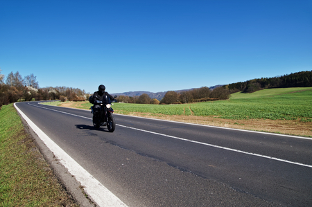Black motorcycle traveling on the asphalt road in rural landscape in early spring. Green fields and flowering trees along the road.の写真素材