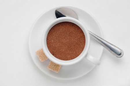 Coffee cup full of ground coffee on saucer with spoon and cane sugar cubes against white background, top view with space for textの写真素材