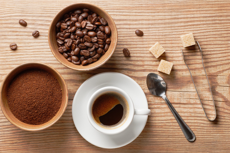 Coffee beans, ground coffee and cup of brewed coffee on rustic wooden table with spoon, sugar tongs, cane sugar cubes and coffee beans, view from aboveの写真素材
