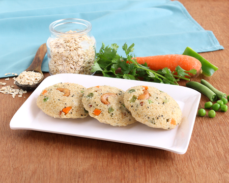 Oats idli or cake, a healthy Indian vegetarian steam-cooked food, with vegetables like carrot and peas, on a ceramic tray.の写真素材