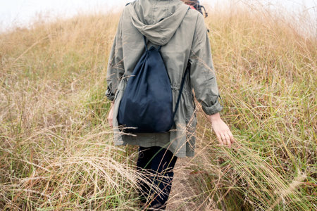 Rear young girl traveler with bag walking to golden meadow on hill
