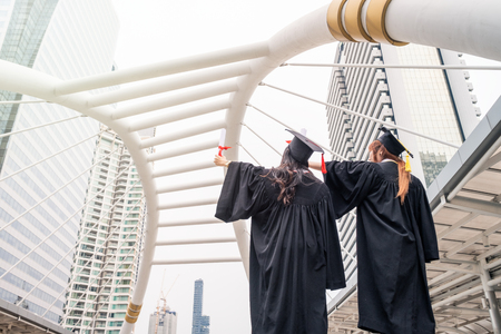 Two young women wearing academic gown raising hand with success Bachelor Degree of Universityの写真素材
