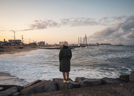 Rear of woman wearing winter coat and hat standing on rocky pier with wave hitting in the sunset at Jumunjin Beach, Gangwon-Do, South Koreaの写真素材