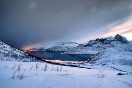 Scenery of snow covered mountain range with illuminated town on coastline in blizzard at mount Segla, Senja island, Norwayの写真素材