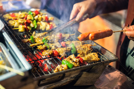 Seller using tongs grilling barbeque pork and sausage on charcoal stove in night marketの写真素材