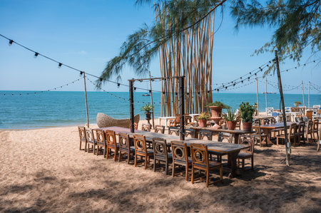 Celebration venue with wooden long table, chair, light bulb hanging and plant decoration in retro style on the beach in tropical seaの写真素材