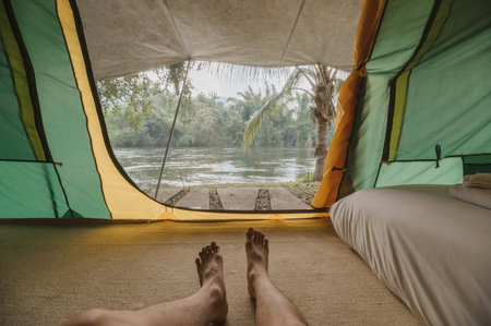 Legs of man relaxing inside a comfortable camping tent on campground in the tropical forest by riverside on summer vacationの写真素材