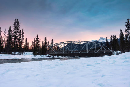 Beautiful sunrise over snowy rocky muntains and the bridge cross the river in pine forest on winter at Banff national park, Alberta, Canadaの写真素材