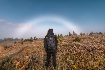 Rear of male traveler adventure standing on golden meadow with fog bow or white rainbow on autumn...