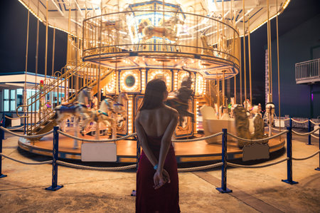 Rear of young asian woman in red dress standing in glowing carousel at amusement parkの写真素材